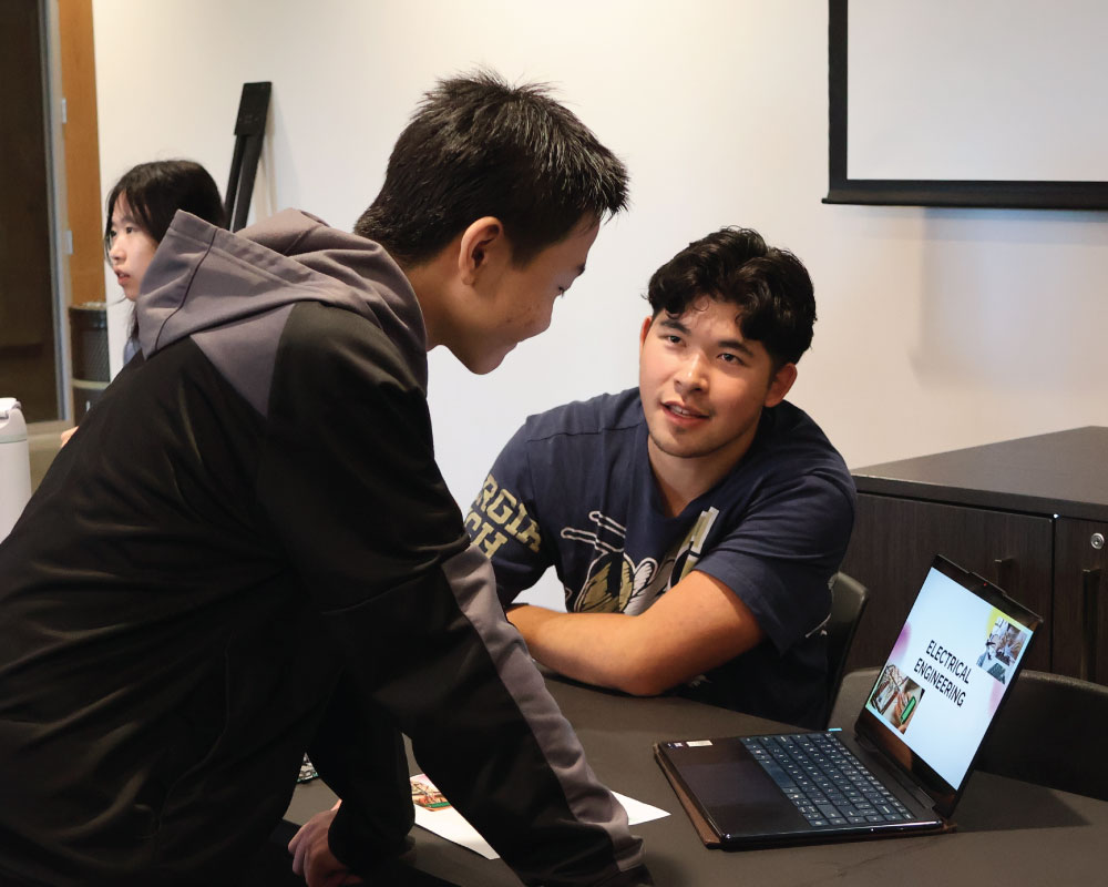 student looking at a laptop while talking to another student